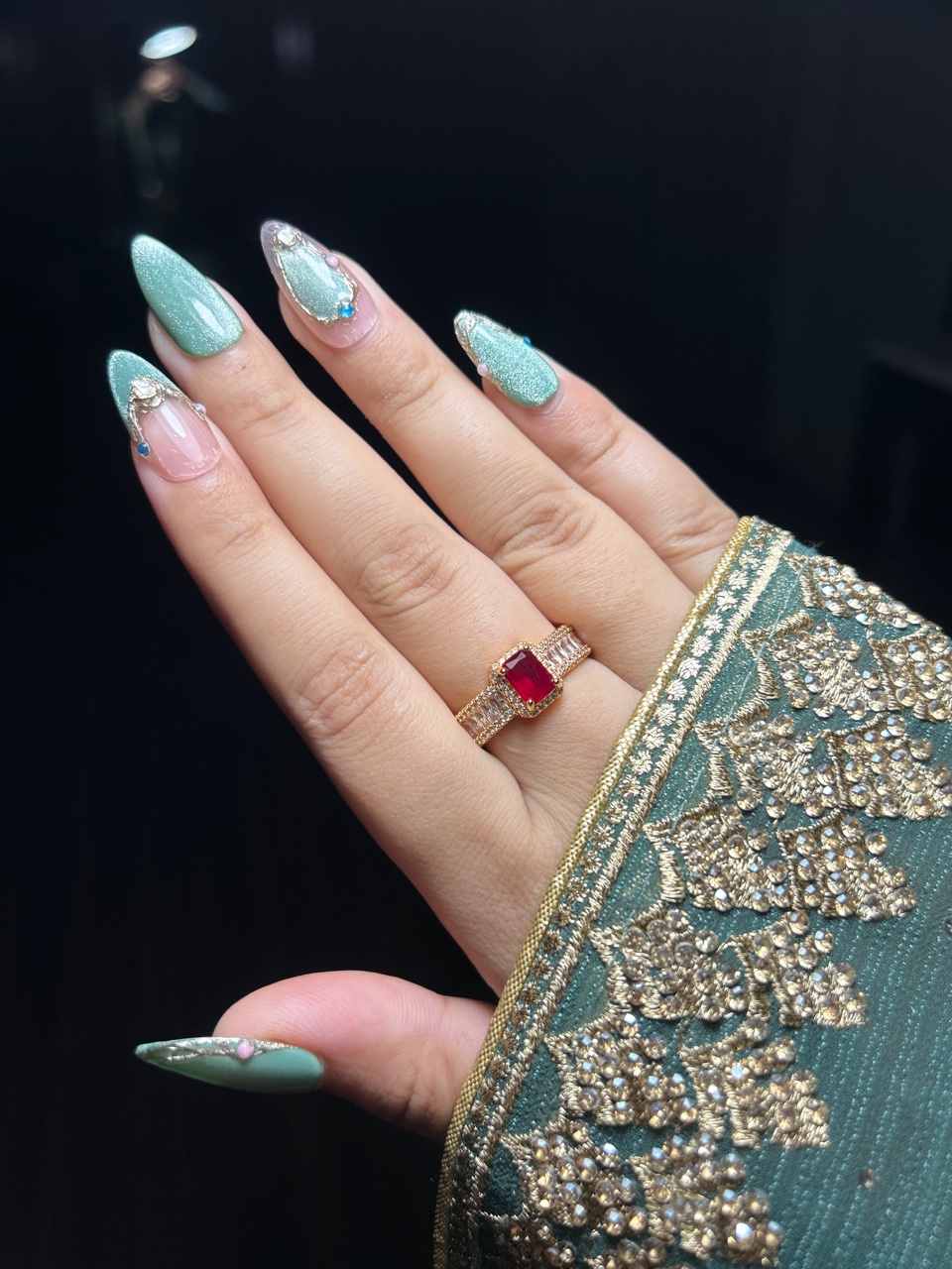 Mint green bridal press-on gel nails with gold French-tip detail and crystal accents, worn with a ruby ring and green-and-gold embroidered outfit, Mehendi night palette