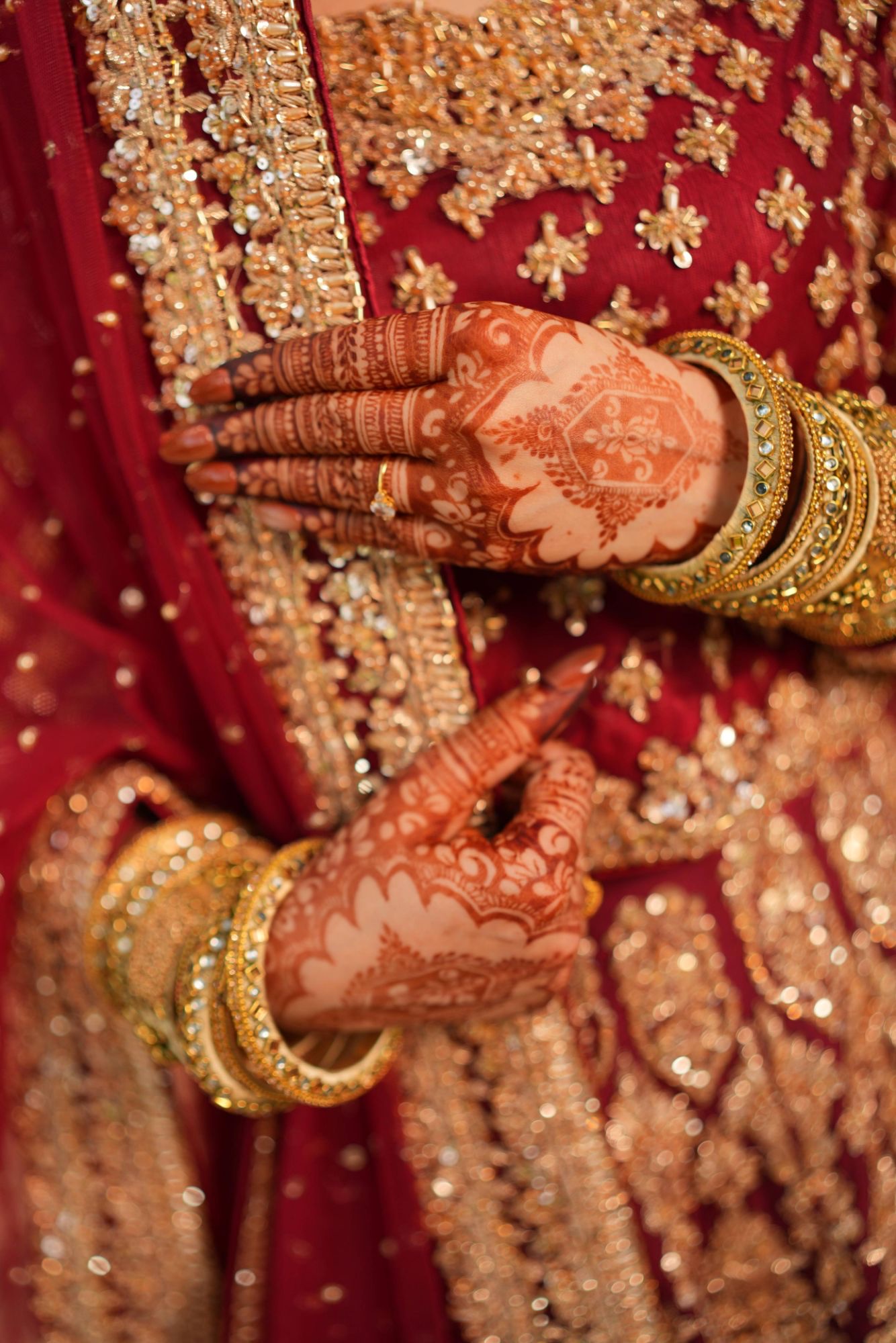 Bride's hennaed hands with long almond press-on nails and gold bangles, resting on a red and gold embroidered bridal dress