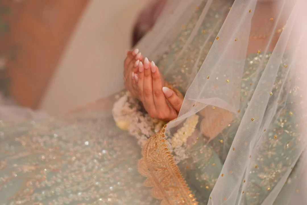 Bride's hands raised in prayer wearing soft nude-to-white ombre almond press-on nails, under a sheer veil over a mint and gold bridal dress