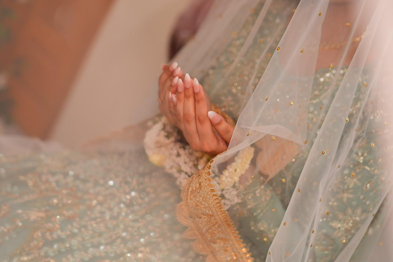 Bride's hands raised in prayer wearing soft nude-to-white ombre almond press-on nails, under a sheer veil over a mint and gold bridal dress