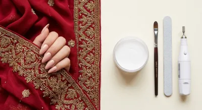 A hand wearing nude-pink almond press-on nails on red and gold bridal fabric, next to salon acrylic tools: acrylic powder, a brush, a nail file and a nail drill