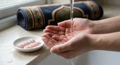 Bare hands cupped under running water for wudu, with a set of press-on nails set aside in a small dish and a rolled prayer mat in the background