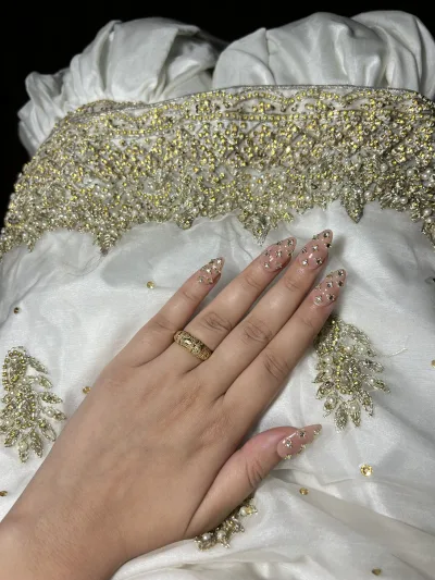 Long almond nude press-on nails with gold beads on a white and gold embroidered dress